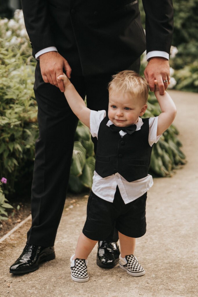 A young child in a black waistcoat, bow tie, white shirt, and chequered slip-on shoes walks on a garden path at Elsham Hall, holding hands with an adult in black formal wear. Lush greenery and small flowers create a perfect scene for any wedding photographer. © Aimee Lince Photography, Lincolnshire photographer