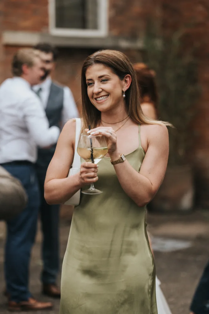 A woman in a light green satin dress holds a glass of white wine, smiling. She has straight brown hair and wears a watch and earrings. Blurred people and a brick wall suggest an outdoor event at Elsham Hall, captured by a wedding photographer. © Aimee Lince Photography, Lincolnshire photographer