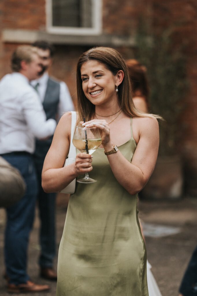 A woman in a light green satin dress holds a glass of white wine, smiling. She has straight brown hair and wears a watch and earrings. Blurred people and a brick wall suggest an outdoor event at Elsham Hall, captured by a wedding photographer. © Aimee Lince Photography, Lincolnshire photographer