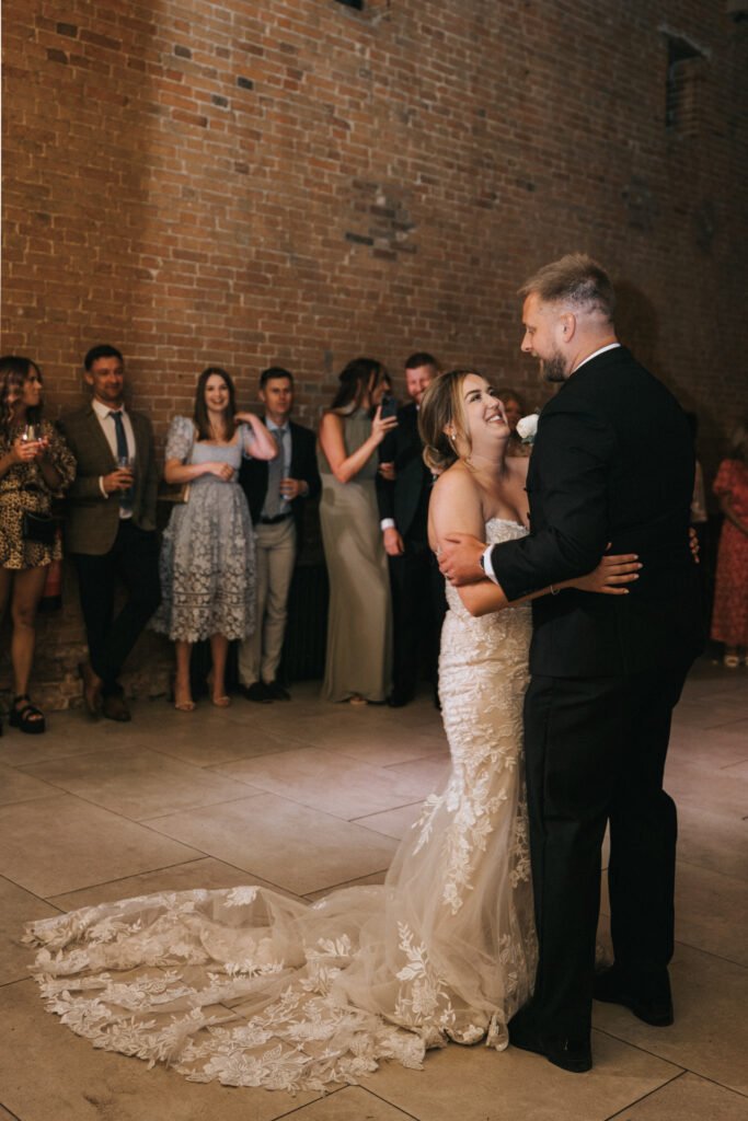 A bride in a strapless, lace wedding dress dances with the groom, who wears a black dinner suit. Captured by their wedding photographer at Elsham Hall, they smile as guests look on and the bride’s train spreads elegantly behind her. © Aimee Lince Photography, Lincolnshire photographer
