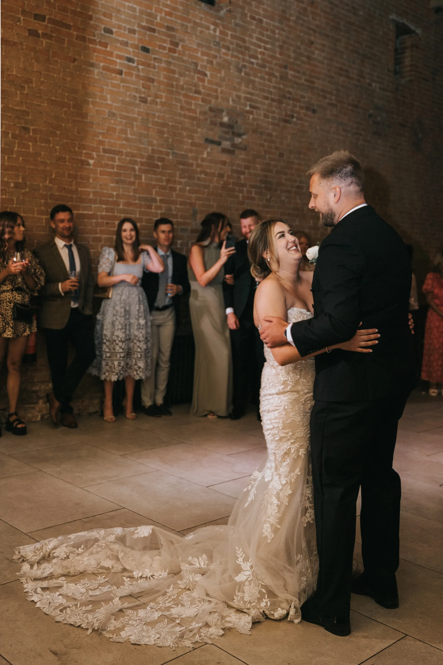 A bride in a strapless, lace wedding dress dances with the groom, who wears a black dinner suit. Captured by their wedding photographer at Elsham Hall, they smile as guests look on and the bride’s train spreads elegantly behind her. © Aimee Lince Photography, Lincolnshire photographer