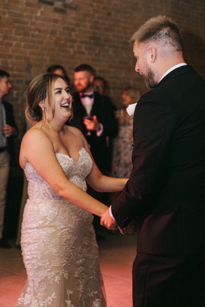 A bride in a strapless lace wedding dress smiles whilst dancing with her groom at Elsham Hall. Captured by a skilled wedding photographer, they hold hands and look at each other, surrounded by guests in formal attire against an exposed brick wall. © Aimee Lince Photography, Lincolnshire photographer