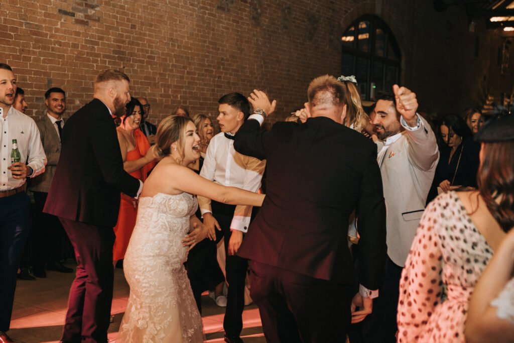 A group of people, including a bride in a white lace dress, joyfully dance together at an indoor Elsham Hall wedding reception. Festive lighting and exposed brick walls set the scene as a wedding photographer captures the lively celebration. © Aimee Lince Photography, Lincolnshire photographer