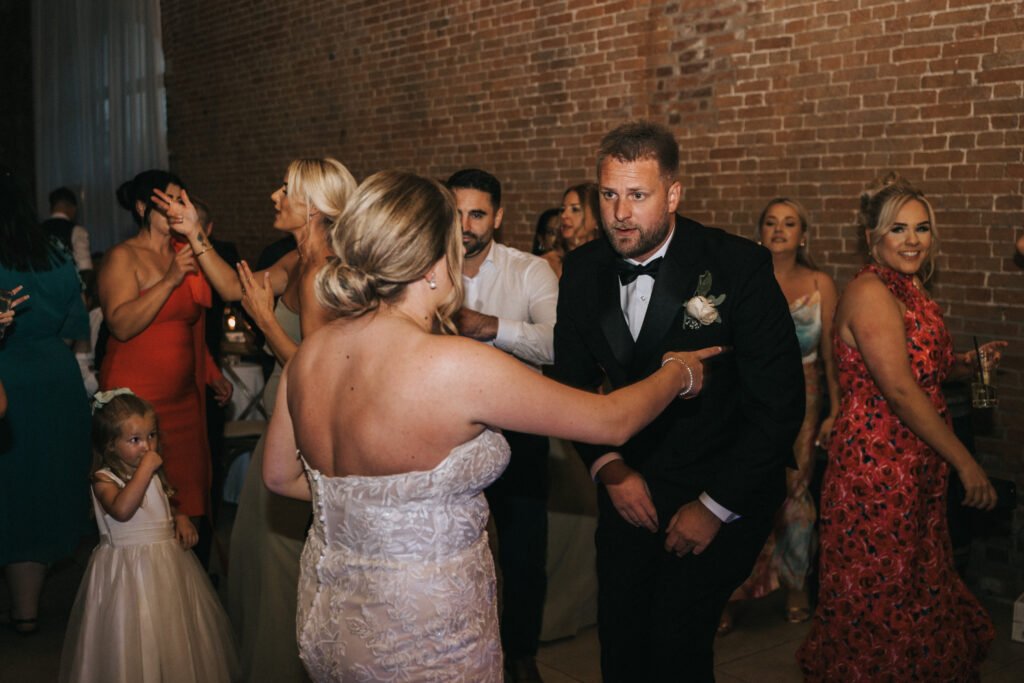 A bride in a strapless lace gown gestures towards a man in a tuxedo with a buttonhole on the dance floor at Elsham Hall. Guests in colourful formal wear and a young girl in white enjoy the moment, perfectly captured by the wedding photographer. © Aimee Lince Photography, Lincolnshire photographer