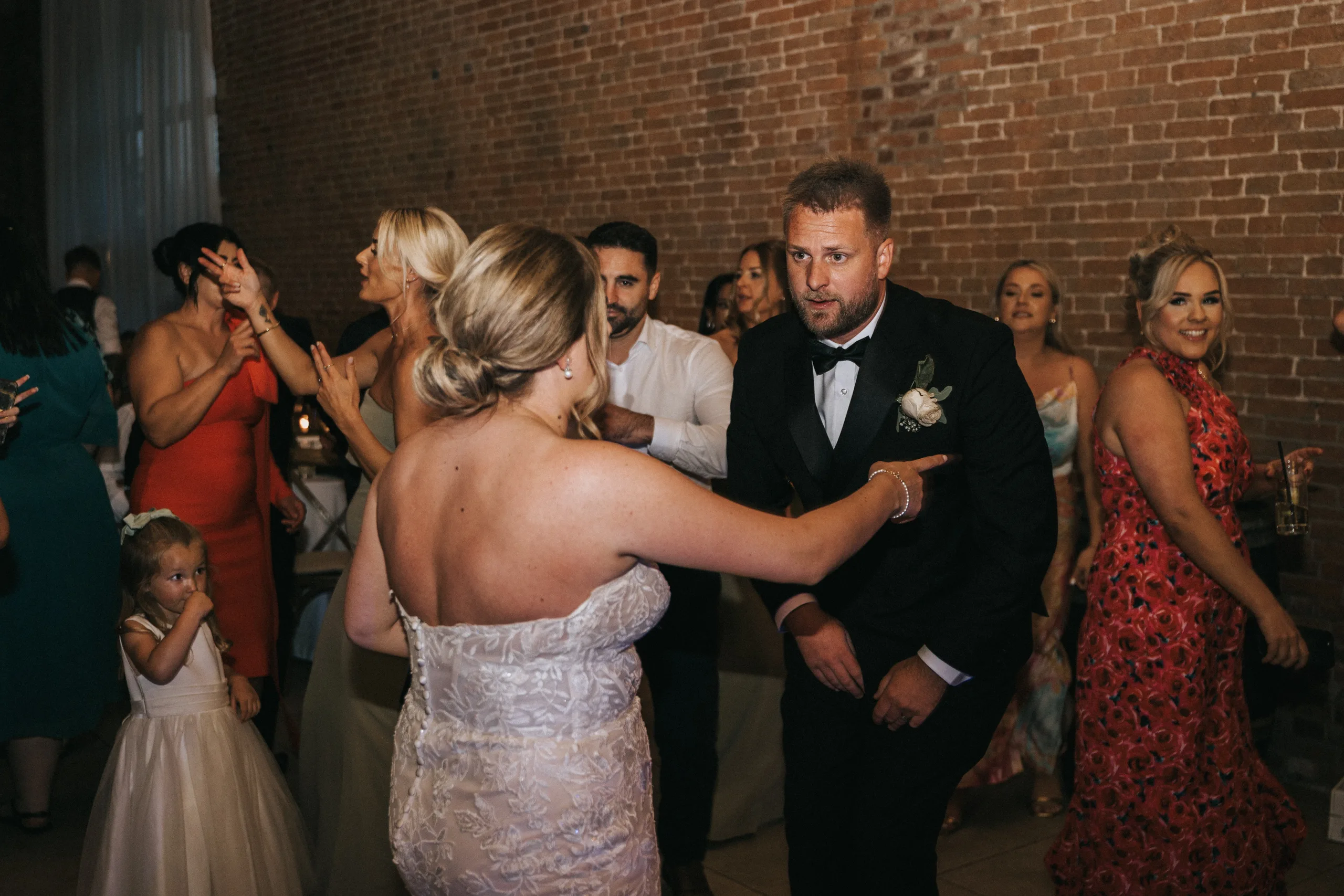 A bride in a strapless lace gown gestures towards a man in a tuxedo with a buttonhole on the dance floor at Elsham Hall. Guests in colourful formal wear and a young girl in white enjoy the moment, perfectly captured by the wedding photographer. © Aimee Lince Photography, Lincolnshire photographer