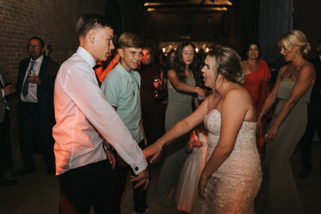 A bride in a strapless white gown energetically dances with a young man in a white shirt. Guests in suits and dresses surround them, some smiling and watching, others dancing, as a wedding photographer captures the joy at rustic Elsham Hall. © Aimee Lince Photography, Lincolnshire photographer