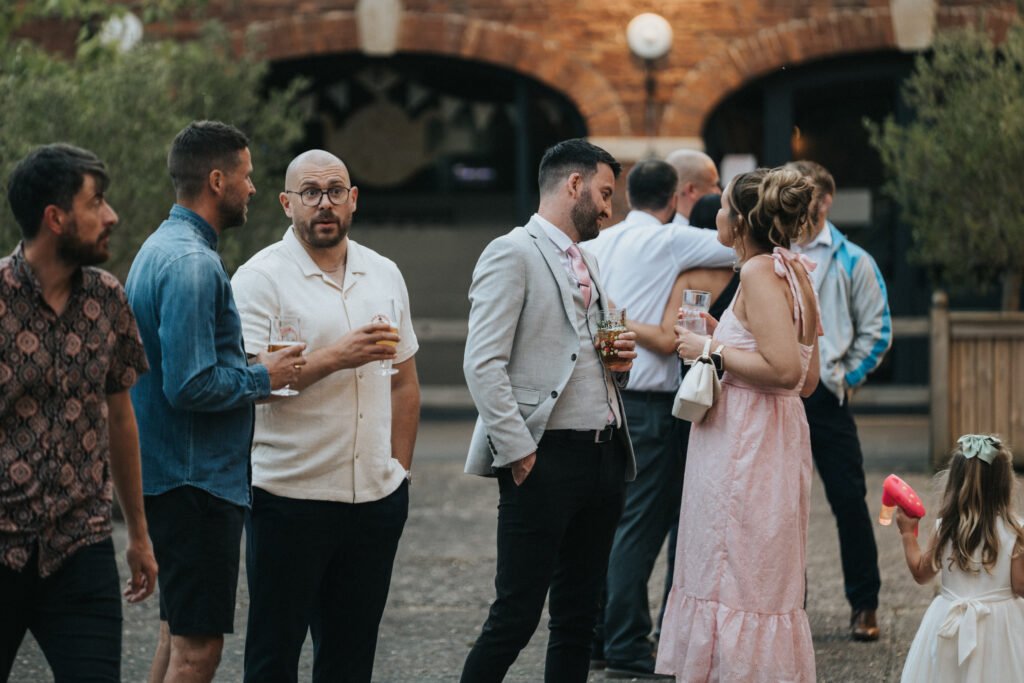 A group of adults in semi-formal attire stand and talk outdoors at Elsham Hall, holding drinks. A young girl in a dress with a pink toy stands to the right. The backdrop, perfect for a wedding photographer, features arched brick doorways and greenery. © Aimee Lince Photography, Lincolnshire photographer