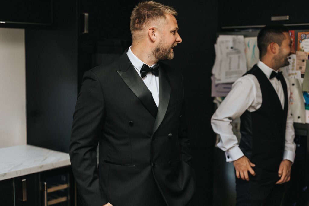 Two men in formalwear stand indoors. The man in the foreground, smiling and looking left, wears a black dinner suit—perhaps captured by a wedding photographer at Elsham Hall. In the background, another man stands by a fridge with colourful drawings. © Aimee Lince Photography, Lincolnshire photographer