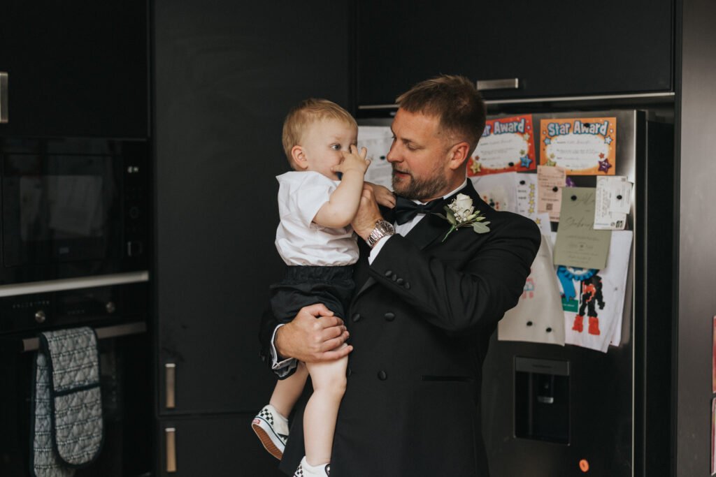 A man in a black suit, like a wedding photographer at Elsham Hall, holds a small child in a kitchen. The man looks at the child, who is wearing a white shirt and shorts. Behind them, the fridge displays colourful drawings, certificates, and magnets. © Aimee Lince Photography, Lincolnshire photographer