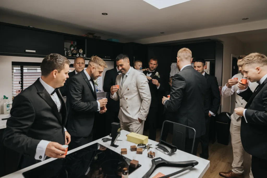 A group of men in suits, some holding drinks, gather and socialise in a modern kitchen at Elsham Hall. One man in a light suit stands out among the others. A wedding photographer captures the scene as a baby is held in the background. © Aimee Lince Photography, Lincolnshire photographer