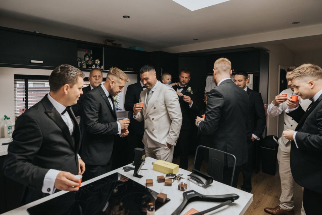 A group of men in suits, some holding drinks, gather and socialise in a modern kitchen at Elsham Hall. One man in a light suit stands out among the others. A wedding photographer captures the scene as a baby is held in the background. © Aimee Lince Photography, Lincolnshire photographer