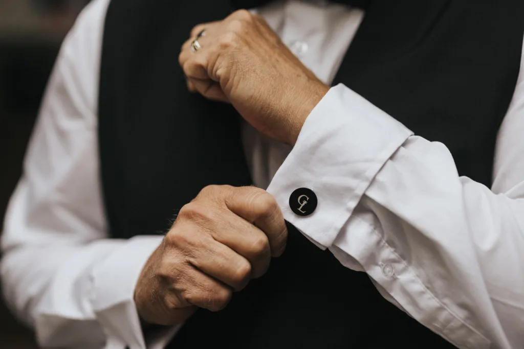 A person in a black waistcoat adjusts their white shirt cuff, showcasing a black cufflink with a monogrammed "G." Their other hand wears a gold ring. The image, by a wedding photographer at Elsham Hall, focuses on the hands and blurred background. © Aimee Lince Photography, Lincolnshire photographer