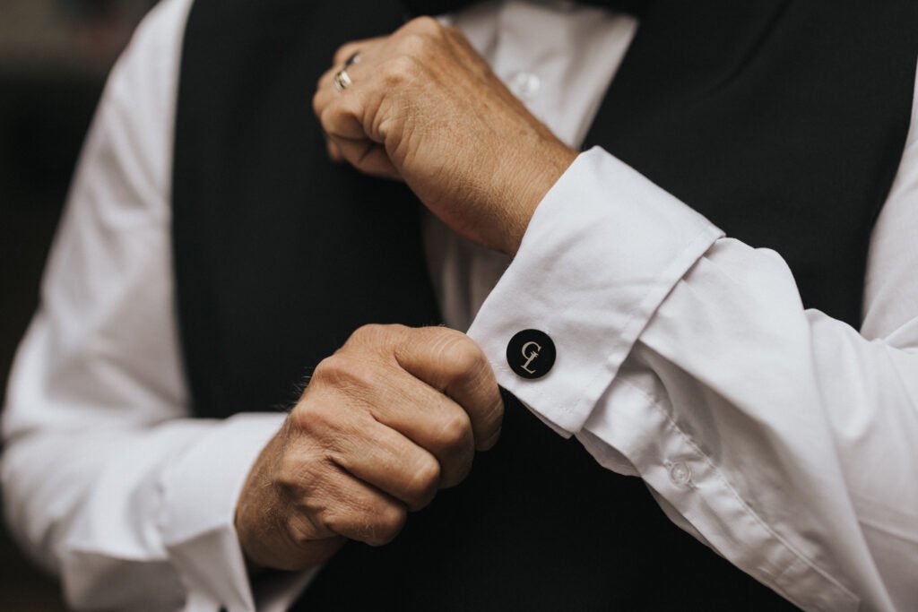 A person in a black waistcoat adjusts their white shirt cuff, showcasing a black cufflink with a monogrammed "G." Their other hand wears a gold ring. The image, by a wedding photographer at Elsham Hall, focuses on the hands and blurred background. © Aimee Lince Photography, Lincolnshire photographer