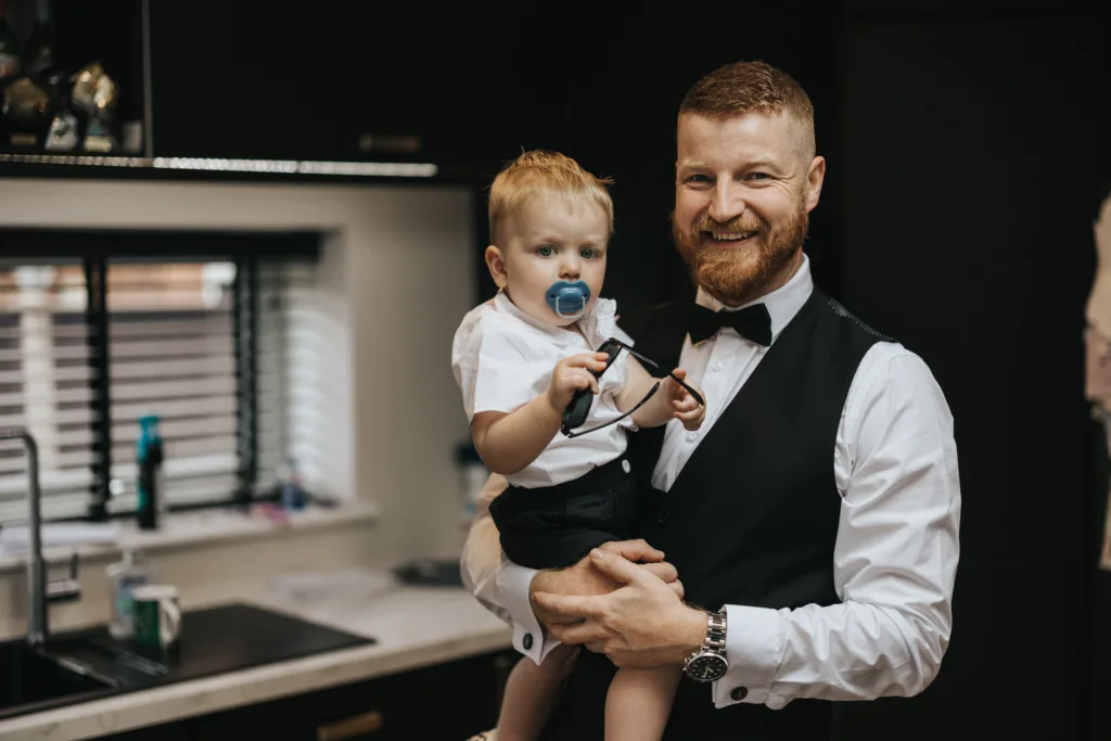 A bearded man in formal attire, possibly a wedding photographer, stands in a modern kitchen holding a smiling child with a dummy. Both wear white shirts and dark shorts, and the man holds a bow tie accessory, ready for an Elsham Hall event. © Aimee Lince Photography, Lincolnshire photographer