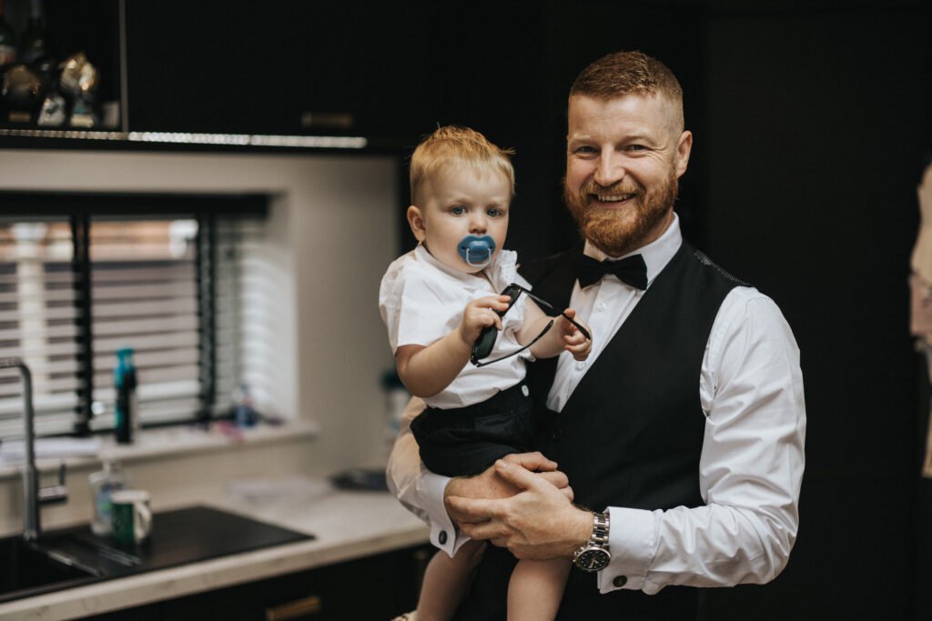 A bearded man in formal attire, possibly a wedding photographer, stands in a modern kitchen holding a smiling child with a dummy. Both wear white shirts and dark shorts, and the man holds a bow tie accessory, ready for an Elsham Hall event. © Aimee Lince Photography, Lincolnshire photographer