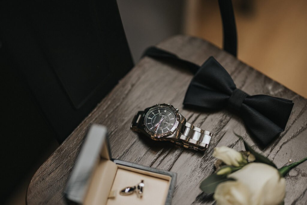 A silver wristwatch, black bow tie, cufflinks in a box, and a white rose buttonhole are arranged neatly on a wooden chair at Elsham Hall, ready for the wedding photographer to capture the elegant, warm atmosphere before the formal event. © Aimee Lince Photography, Lincolnshire photographer