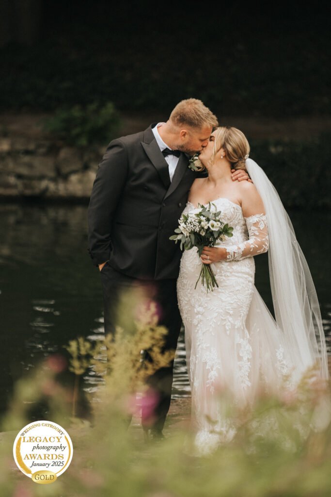 A bride and groom share a kiss outdoors by a pond at Elsham Hall. The groom wears a black suit; the bride is in white lace, holding a bouquet. Lush greenery surrounds them. An award badge marks the image by their wedding photographer in the lower left corner. © Aimee Lince Photography, Lincolnshire photographer