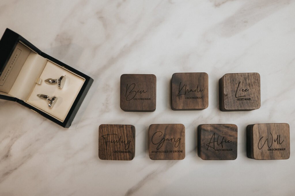 Seven engraved wooden boxes are arranged on a marble surface at Elsham Hall, each labelled with a name and wedding role. To the left, an open black box displays shiny silver cufflinks—perfect details for a wedding photographer to capture. © Aimee Lince Photography, Lincolnshire photographer
