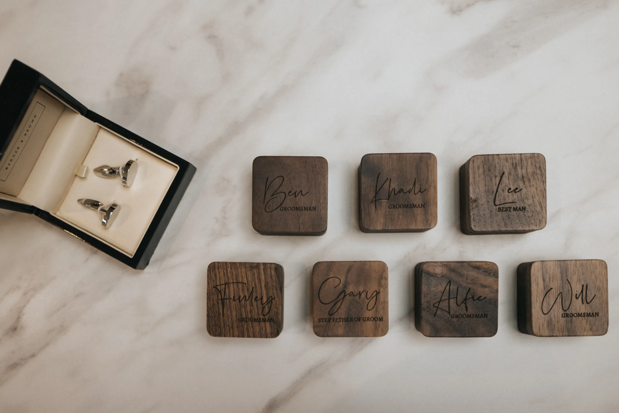 Seven engraved wooden boxes are arranged on a marble surface at Elsham Hall, each labelled with a name and wedding role. To the left, an open black box displays shiny silver cufflinks—perfect details for a wedding photographer to capture. © Aimee Lince Photography, Lincolnshire photographer