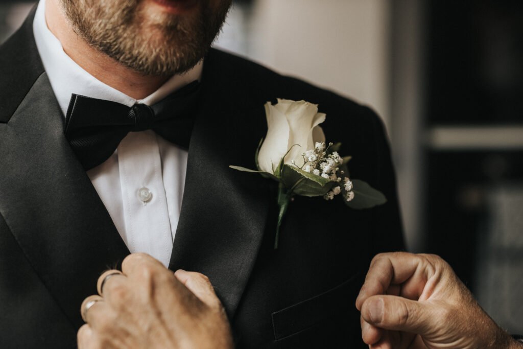 A close-up of a man in a black dinner jacket and bow tie, with a white rose buttonhole being pinned to his lapel by another’s hands—captured by a wedding photographer at Elsham Hall. The man has a short beard and moustache; the background is softly blurred. © Aimee Lince Photography, Lincolnshire photographer