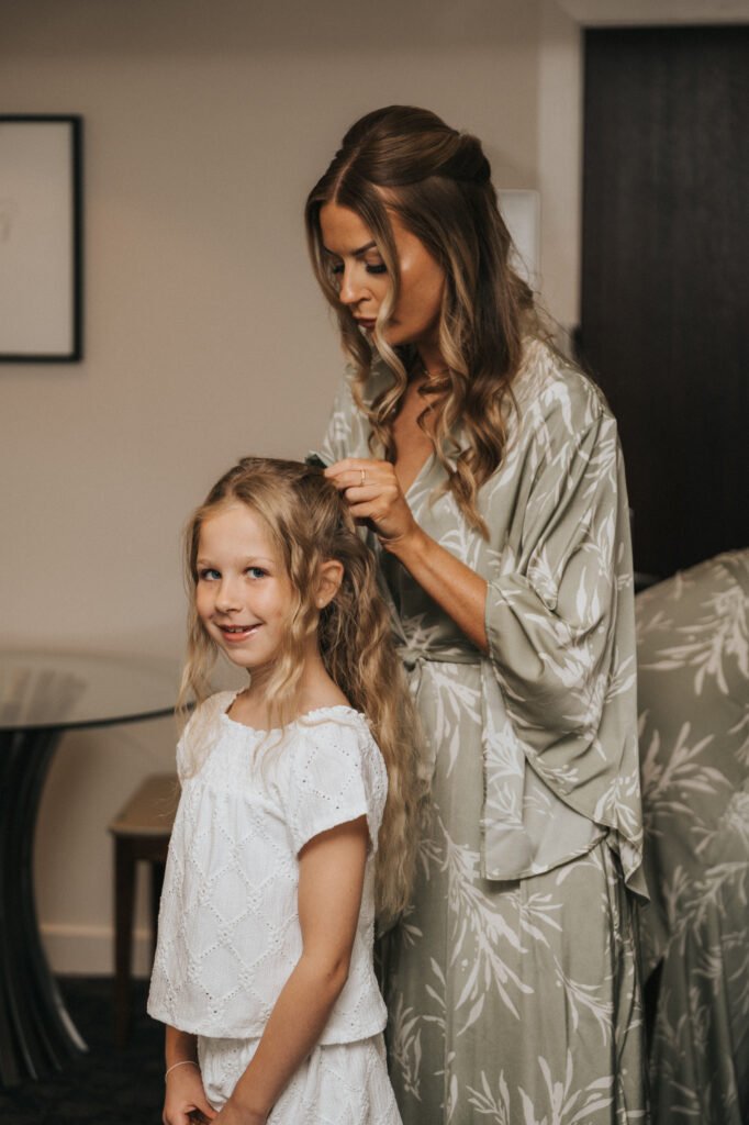A woman in a light green floral dressing gown styles the hair of a smiling young girl with long blonde hair and a white broderie anglaise top. Indoors, against neutral decor, they share a sweet moment—perfect for an Elsham Hall wedding photographer to capture. © Aimee Lince Photography, Lincolnshire photographer