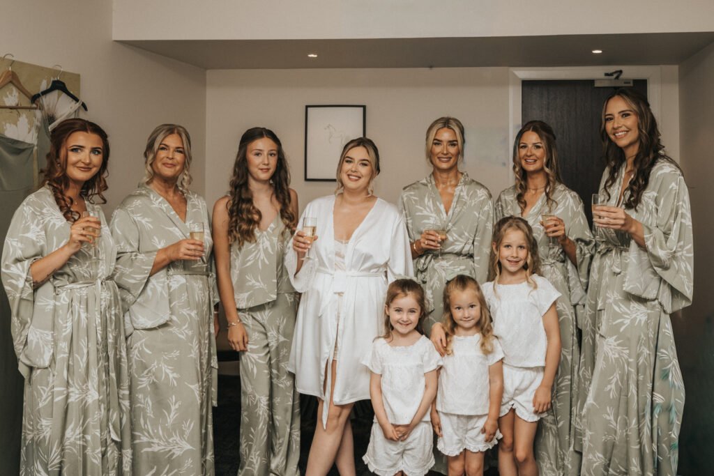 Eight women in matching sage green dressing gowns, except one in white, smile and hold drinks in a cosy, modern room at Elsham Hall. Four young girls in white outfits stand in front, also smiling—captured beautifully by their wedding photographer. © Aimee Lince Photography, Lincolnshire photographer