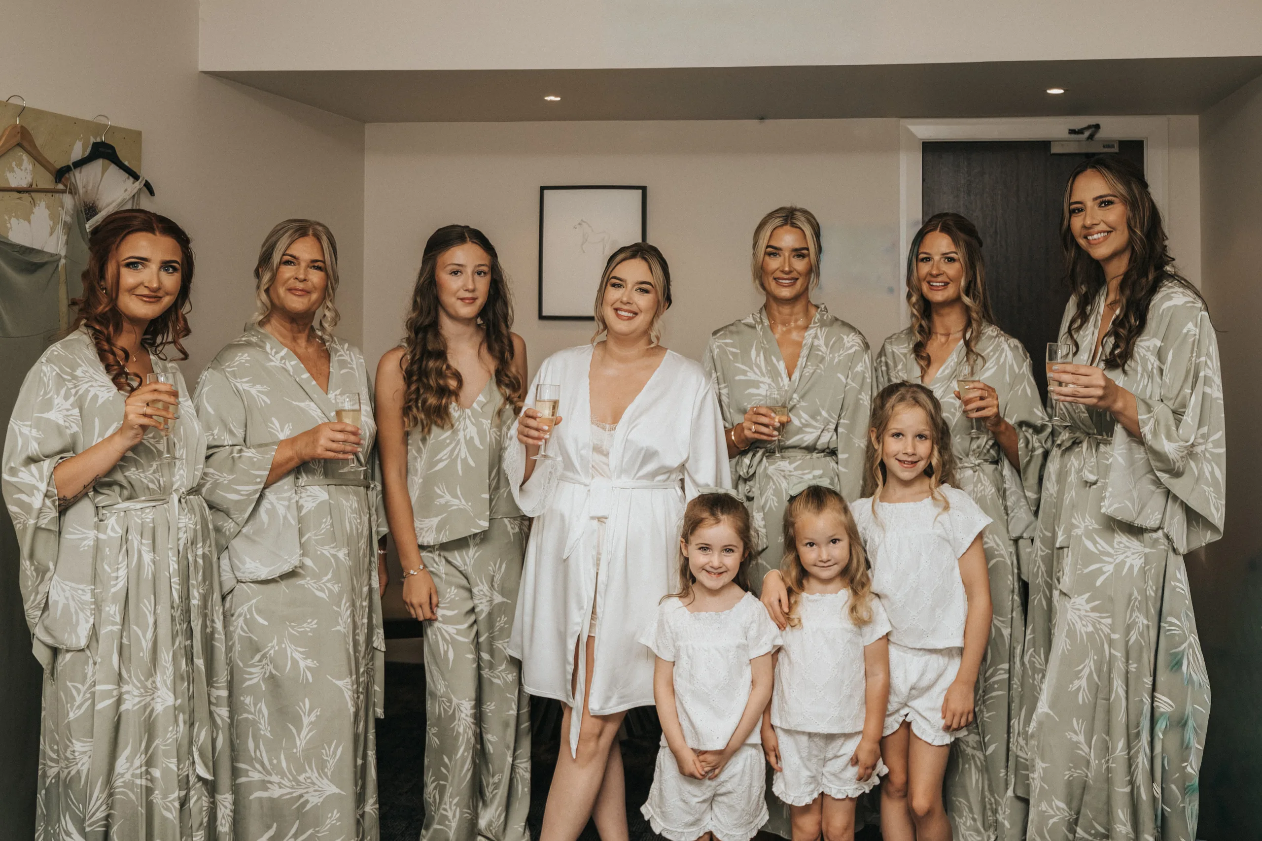 Eight women in matching sage green dressing gowns, except one in white, smile and hold drinks in a cosy, modern room at Elsham Hall. Four young girls in white outfits stand in front, also smiling—captured beautifully by their wedding photographer. © Aimee Lince Photography, Lincolnshire photographer