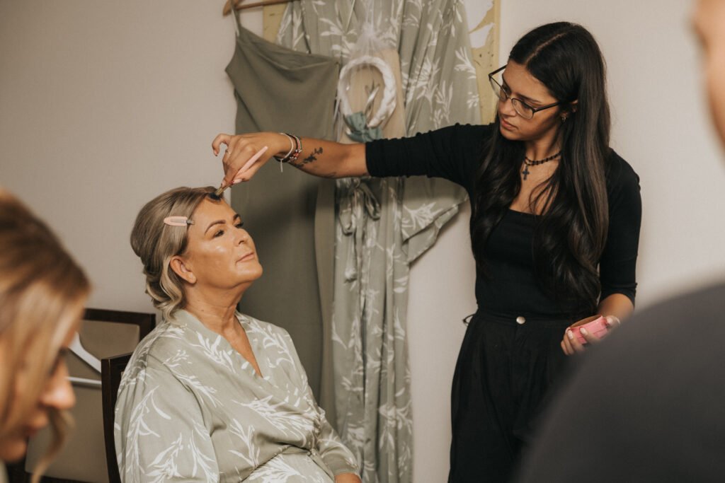 A woman with fair hair sits in a dressing gown, eyes closed, as a wedding photographer captures the calm moment while a make-up artist applies make-up to her forehead. A green dress hangs in the background at Elsham Hall, adding to the tranquil setting. © Aimee Lince Photography, Lincolnshire photographer