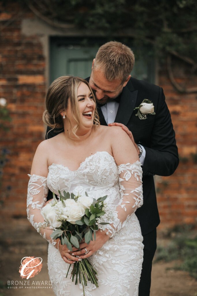 A joyful bride in a white lace off-shoulder gown holds a bouquet of white roses while laughing. Her groom, smiling, leans in behind her as they pose outdoors against a rustic brick wall at Elsham Hall—perfect for any wedding photographer to capture. © Aimee Lince Photography, Lincolnshire photographer
