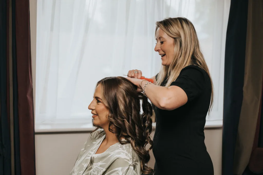 A hairdresser in a black outfit smiles as she styles the long, wavy hair of a seated woman in a satin dressing gown. Indoors by sheer white curtains, they prepare for the day—a moment perfectly suited for an Elsham Hall wedding photographer to capture. © Aimee Lince Photography, Lincolnshire photographer