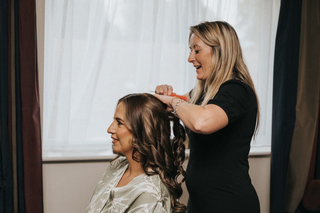 A hairdresser in a black outfit smiles as she styles the long, wavy hair of a seated woman in a satin dressing gown. Indoors by sheer white curtains, they prepare for the day—a moment perfectly suited for an Elsham Hall wedding photographer to capture. © Aimee Lince Photography, Lincolnshire photographer