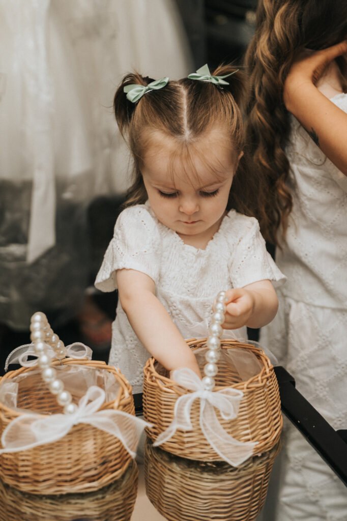A young girl with light brown hair in plaits and green bows, captured by a wedding photographer at Elsham Hall, wears a white dress as she places her hand inside one of two wicker baskets decorated with ribbon and pearl handles. Another child is partially visible beside her. © Aimee Lince Photography, Lincolnshire photographer