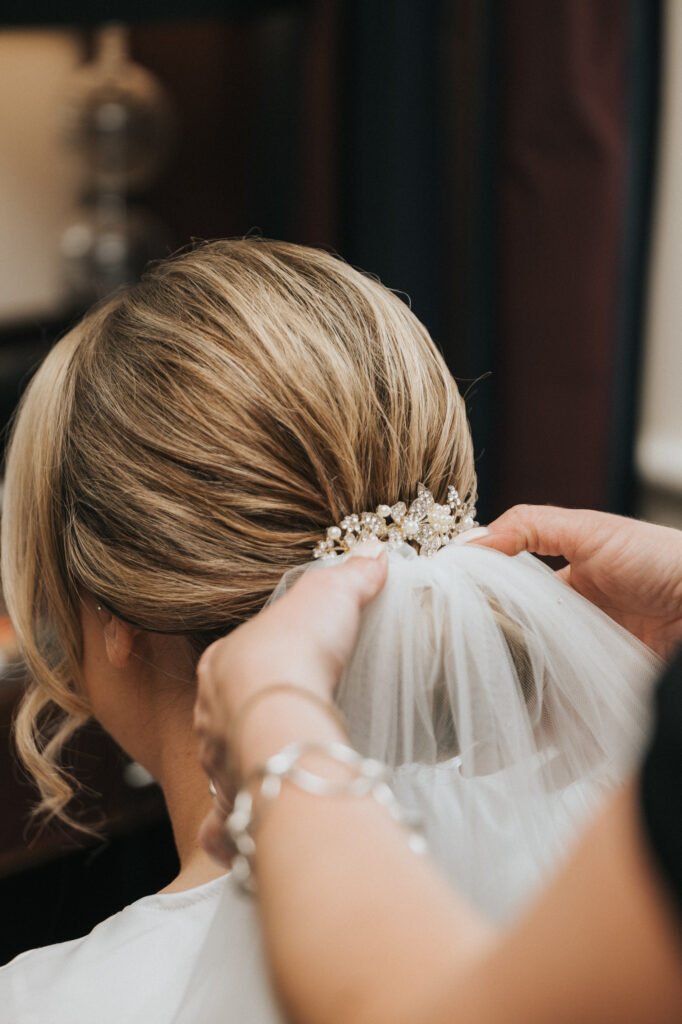 A close-up of a bride’s blonde hair in an elegant low bun as someone fastens a sparkling jewelled hairpiece and veil. Captured by a wedding photographer at Elsham Hall, her softly curled hair is perfectly framed indoors with her face turned away. © Aimee Lince Photography, Lincolnshire photographer
