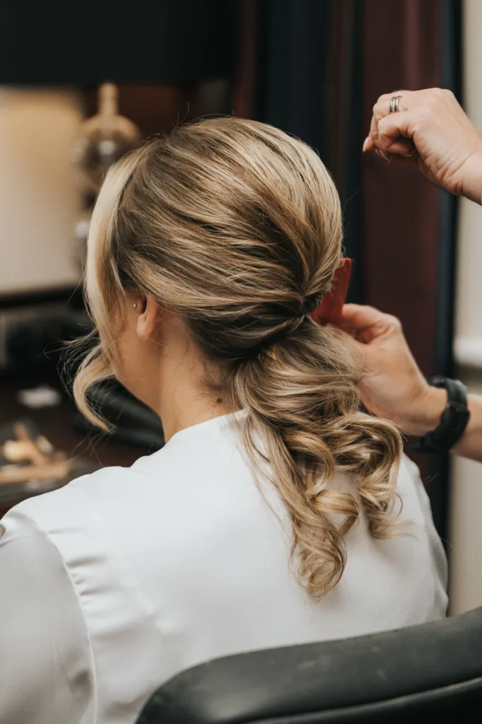 A person with blonde, curled hair styled in a low, elegant updo sits facing away as a wedding photographer captures someone adjusting the hairstyle with a red comb. The individual wears a white dressing gown, set against softly blurred indoor furnishings. © Aimee Lince Photography, Lincolnshire photographer