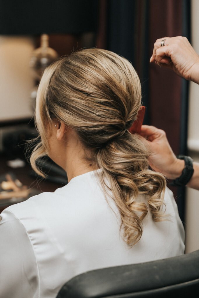 A person with blonde, curled hair styled in a low, elegant updo sits facing away as a wedding photographer captures someone adjusting the hairstyle with a red comb. The individual wears a white dressing gown, set against softly blurred indoor furnishings. © Aimee Lince Photography, Lincolnshire photographer