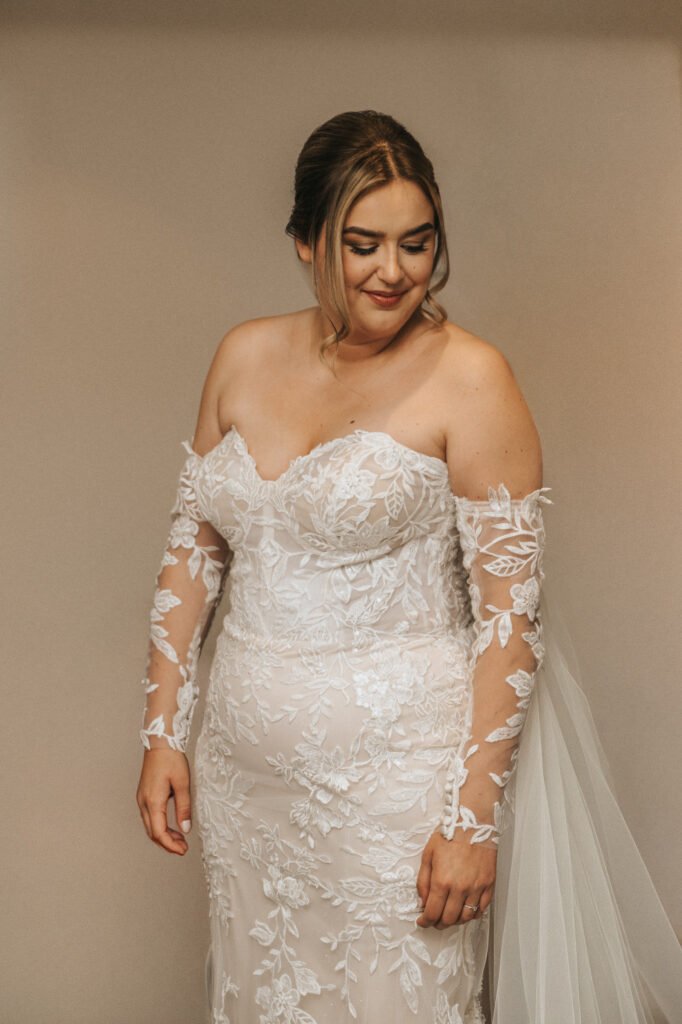 A bride stands against a plain background, wearing an off-the-shoulder, white lace wedding dress with floral patterns. Captured by a wedding photographer at Elsham Hall, her hair is styled in an elegant up-do as she looks down slightly, smiling softly. © Aimee Lince Photography, Lincolnshire photographer