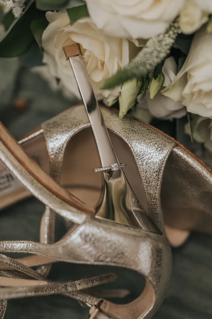 A diamond engagement ring rests on the metallic heel of a gold high-heeled shoe at Elsham Hall, surrounded by white roses and greenery, creating an elegant and romantic composition perfect for a wedding photographer. © Aimee Lince Photography, Lincolnshire photographer