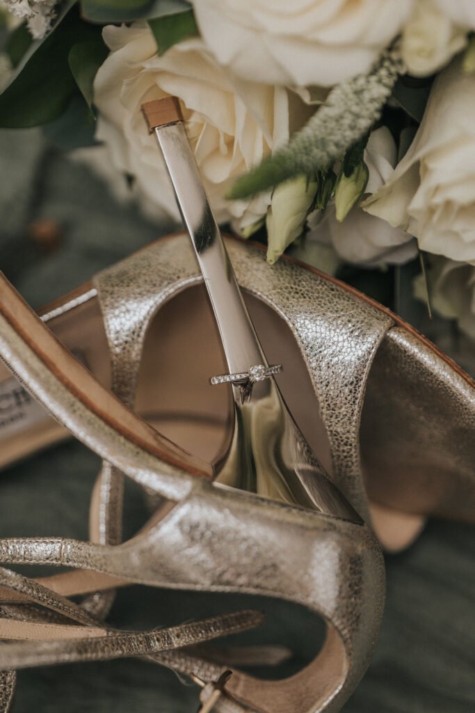 A diamond engagement ring rests on the metallic heel of a gold high-heeled shoe at Elsham Hall, surrounded by white roses and greenery, creating an elegant and romantic composition perfect for a wedding photographer. © Aimee Lince Photography, Lincolnshire photographer