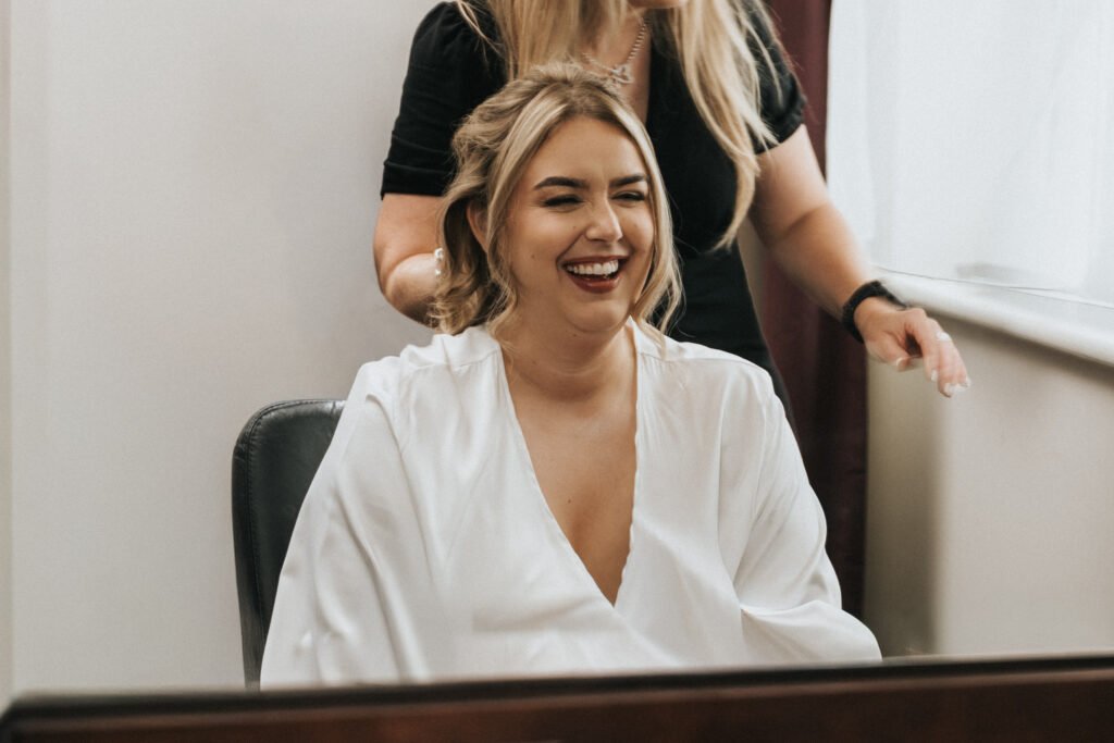 A woman in a white dressing gown sits smiling in front of a mirror at Elsham Hall, while another person with long blonde hair styles her hair—an ideal moment for a wedding photographer to capture the joy and natural light streaming through the window. © Aimee Lince Photography, Lincolnshire photographer