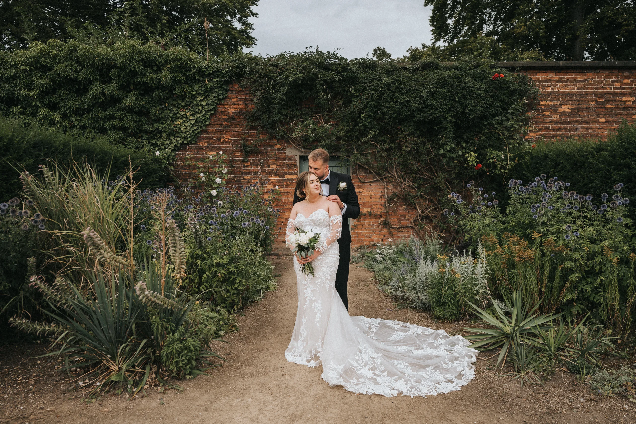 A bride in a white, off-shoulder gown holding a bouquet stands on a garden path at Elsham Hall, whilst her groom lovingly embraces her from behind. Lush green plants and ivy-covered walls frame this timeless moment, perfect for any wedding photographer. © Aimee Lince Photography, Lincolnshire photographer