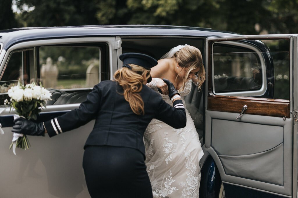 A wedding photographer captures a bride in a white dress and veil as she is helped out of a classic car by a chauffeur. He holds her bouquet amid greenery and blurred gravestones, creating a memorable scene at Elsham Hall. © Aimee Lince Photography, Lincolnshire photographer