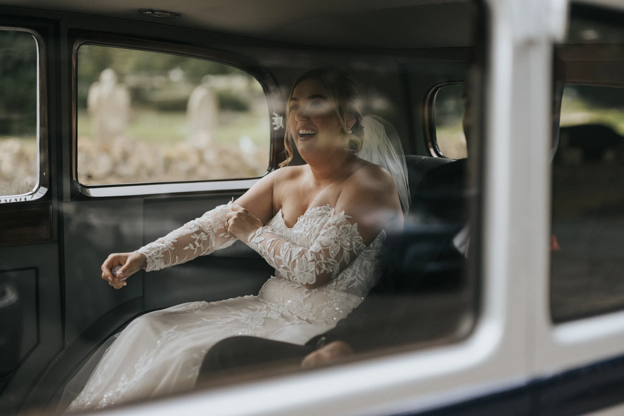 A joyful bride in an off-the-shoulder, lace wedding dress and veil sits inside a vintage car, smiling and laughing. Captured by a wedding photographer at Elsham Hall, the image shows greenery outside as she gestures with delight through the window. © Aimee Lince Photography, Lincolnshire photographer