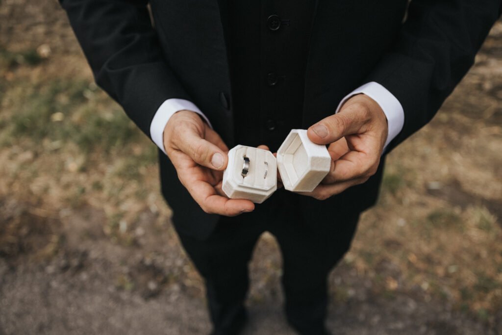 A wedding photographer captures a person in a black suit holding an open white ring box with two rings, standing on a grassy dirt path—hands, torso, and part of the suit visible—at Elsham Hall. © Aimee Lince Photography, Lincolnshire photographer