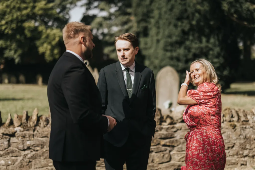 Three adults stand outdoors by a stone wall at Elsham Hall, engaged in conversation. Two men wear dark suits and ties, whilst a woman in a bright pink floral dress smiles, brushing hair from her face—perfect for a wedding photographer to capture. © Aimee Lince Photography, Lincolnshire photographer
