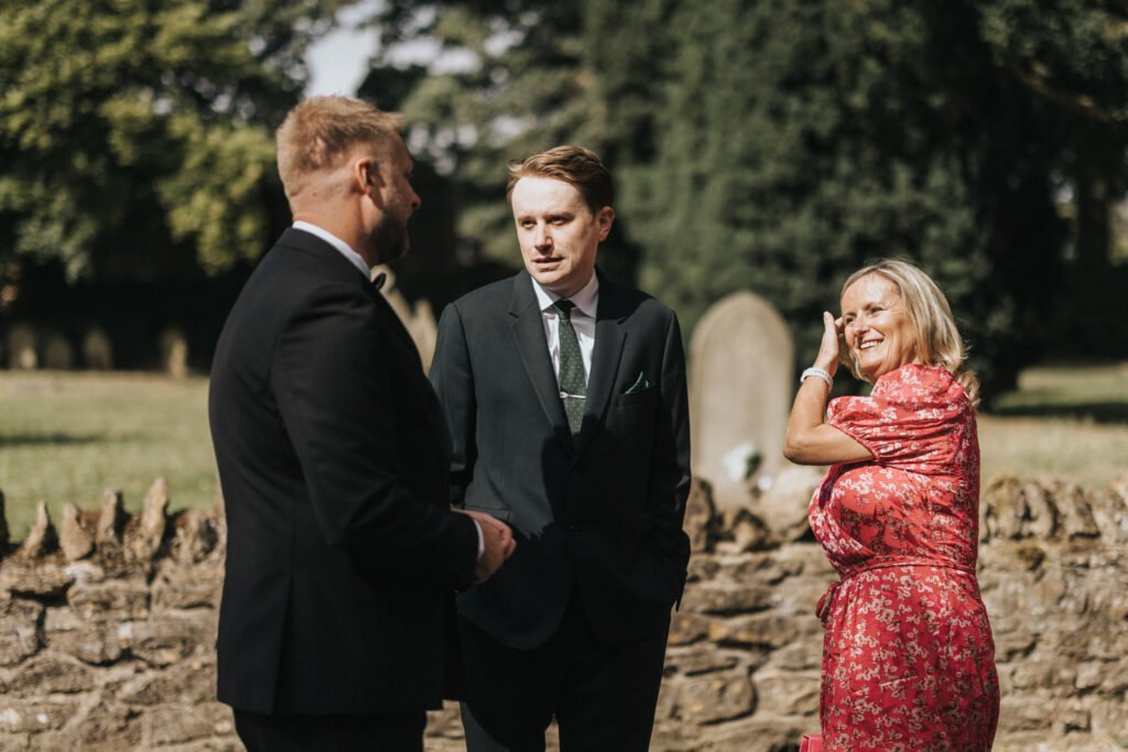 Three adults stand outdoors by a stone wall at Elsham Hall, engaged in conversation. Two men wear dark suits and ties, whilst a woman in a bright pink floral dress smiles, brushing hair from her face—perfect for a wedding photographer to capture. © Aimee Lince Photography, Lincolnshire photographer