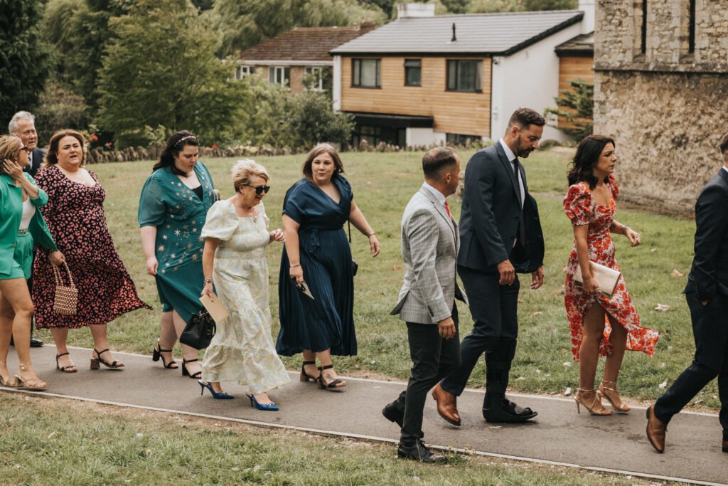 A group of well-dressed people, including women in dresses and men in suits, walk together at Elsham Hall on a paved path near grass, trees, and buildings. The festive atmosphere suggests they are attending a celebration captured by a wedding photographer. © Aimee Lince Photography, Lincolnshire photographer