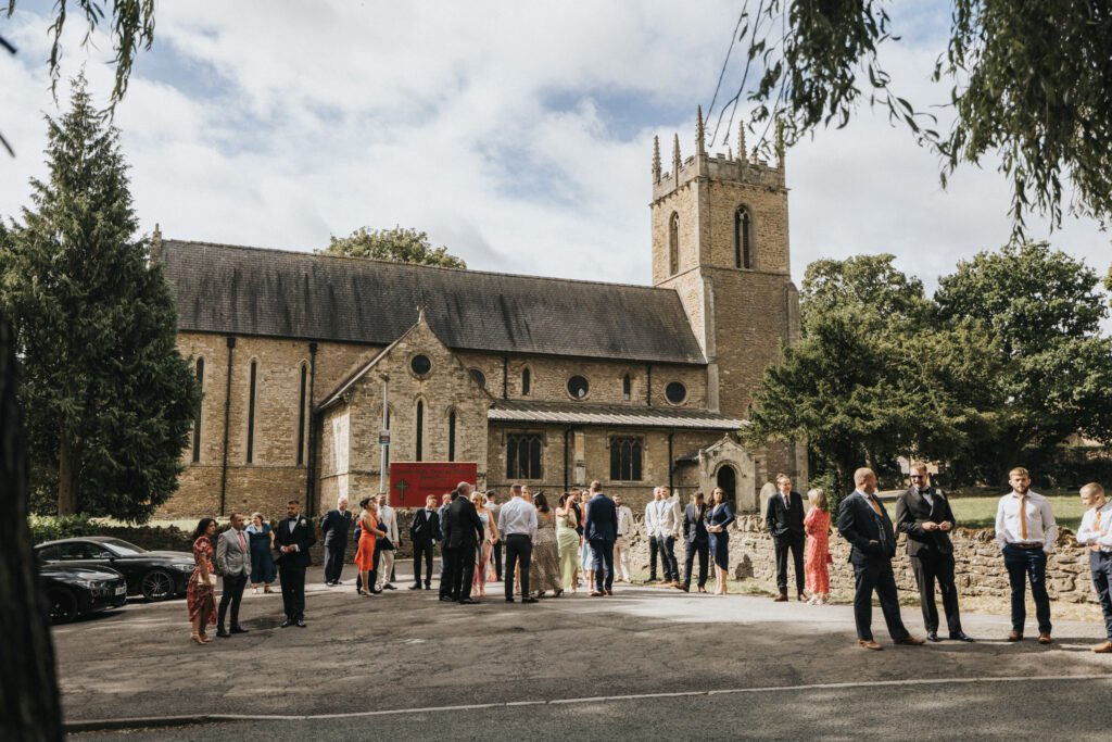 A group of people dressed formally gather outside a stone church with arched windows and a tall clock tower on a sunny day. Trees frame the scene, and cars are parked nearby—a festive mood, perfect for a wedding photographer or Elsham Hall celebration. © Aimee Lince Photography, Lincolnshire photographer