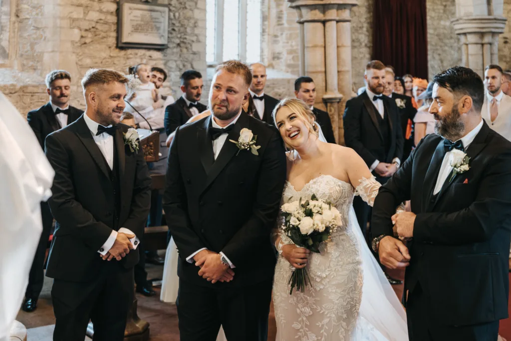 A bride in a white gown and three men in black dinner jackets stand at the front of a stone church. Captured by a wedding photographer, light streams through arched windows as guests watch from the pews, evoking the timeless beauty of Elsham Hall. © Aimee Lince Photography, Lincolnshire photographer