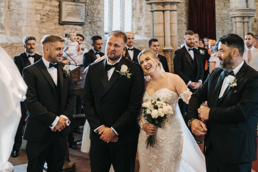 A bride in a white gown and three men in black dinner jackets stand at the front of a stone church. Captured by a wedding photographer, light streams through arched windows as guests watch from the pews, evoking the timeless beauty of Elsham Hall. © Aimee Lince Photography, Lincolnshire photographer