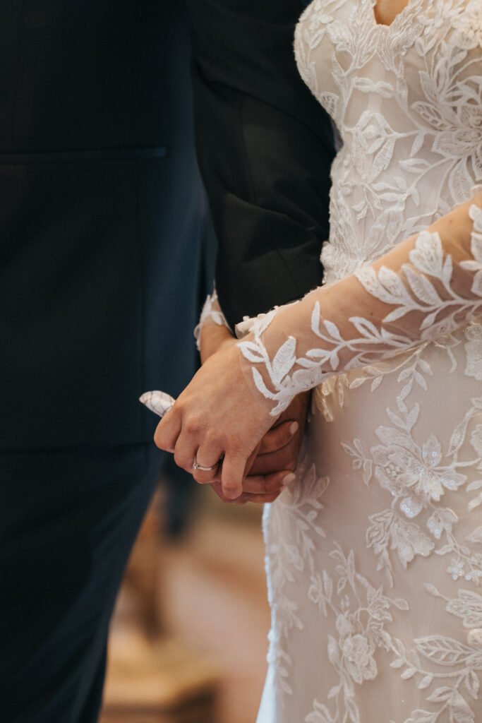 A bride in an elegant, long-sleeved lace wedding dress holds hands with a person in a dark suit. Captured by a wedding photographer at Elsham Hall, the focus is on their joined hands, her wedding ring, and the delicate floral lace details. © Aimee Lince Photography, Lincolnshire photographer